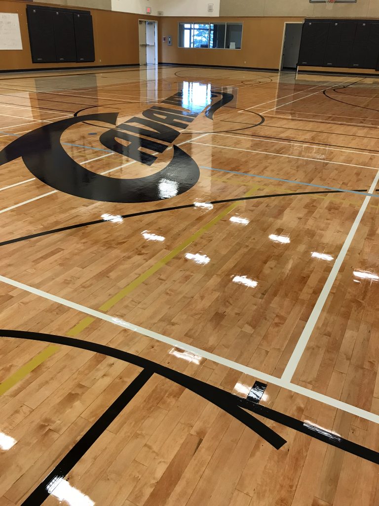 wa“Indoor basketball court with glossy wooden floor and white boundary lines, featuring a basketball hoop at the far end against a mural-adorned wall, with banners and signs on the walls, and an orange basketball near the hoop.”

Description: The court has a polished wooden floor with a reflective surface that catches the overhead lights. The playing area is marked with white lines, and there’s a large green area within the three-point arc. At the end of the court, there’s a basketball hoop set against a wall that has murals or designs, which add a decorative touch to the space. The walls are also embellished with banners or signs, which could signify achievements or affiliations with sports teams or organizations. The court is currently empty, with no players in sight, and there’s a single orange basketball positioned near the hoop. This setting could be ideal for sports events, basketball practice, or as a facility highlight for a venue promotion.ht can i say for alt text this image