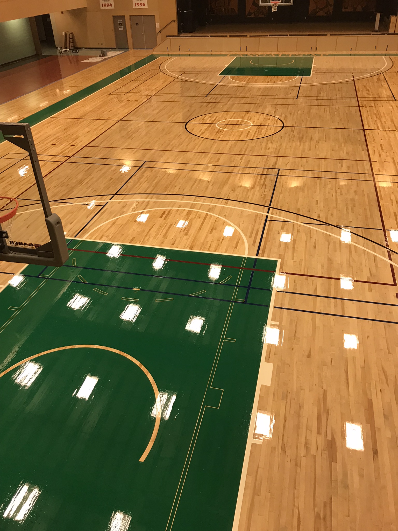 Indoor basketball court with glossy wooden floor and white boundary lines, featuring a basketball hoop at the far end against a mural-adorned wall, with banners and signs on the walls, and an orange basketball near the hoop