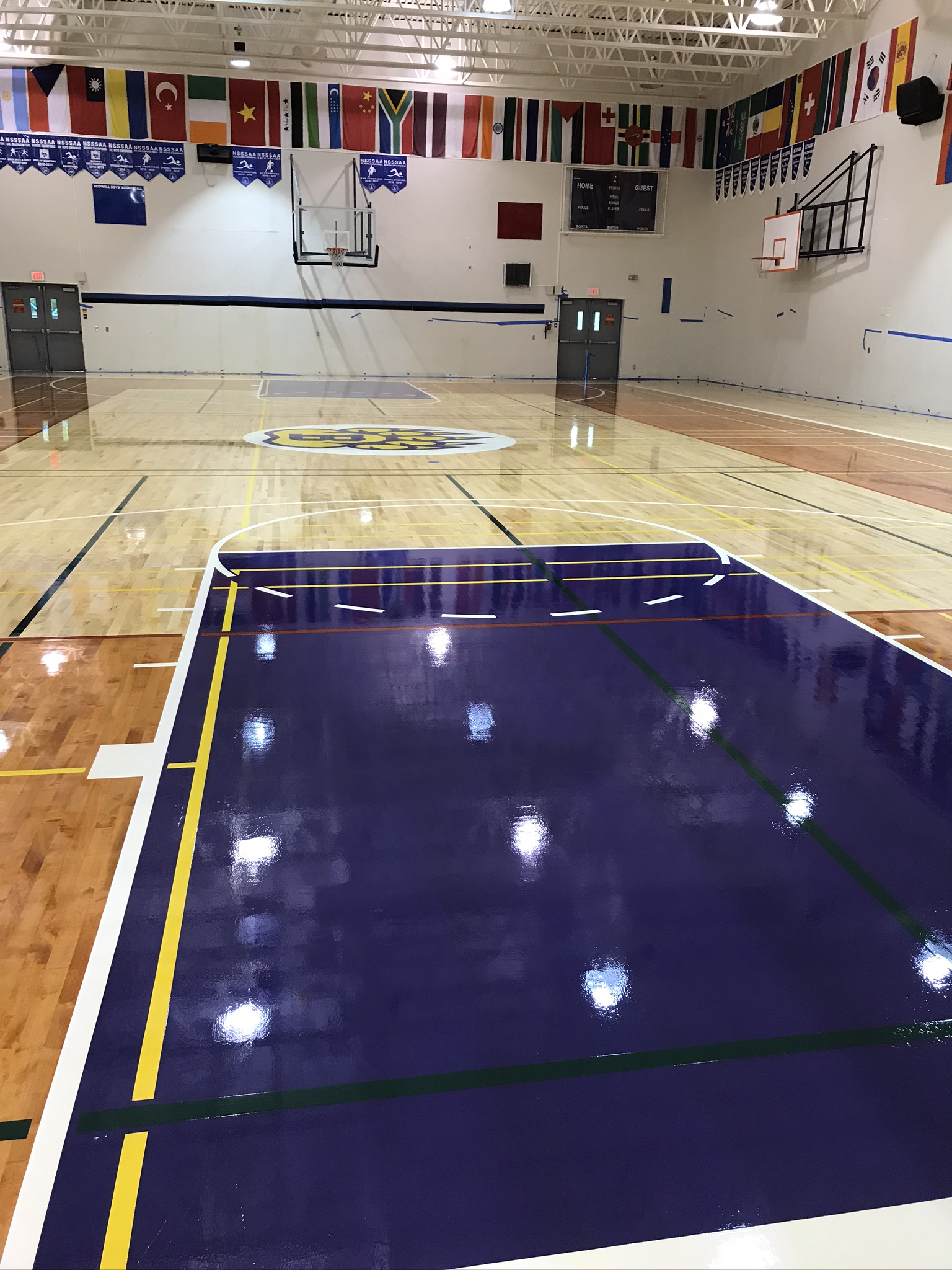 he image you’ve provided appears to be of an indoor basketball court with a polished wooden floor that reflects the overhead lights. The court is marked with white and blue lines indicating the boundaries, the center circle, free throw lanes, and the three-point lines. There are two basketball hoops visible, one at each end of the court. The green painted areas denote the key or paint areas under each basket. This setting is typical for sports facilities, basketball games, or athletic training environments.